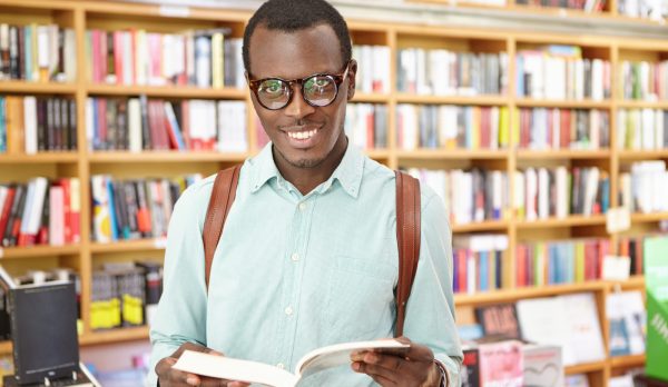 cheerful-young-fashionable-black-man-wearing-glasses-standing-library-with-shelves-books