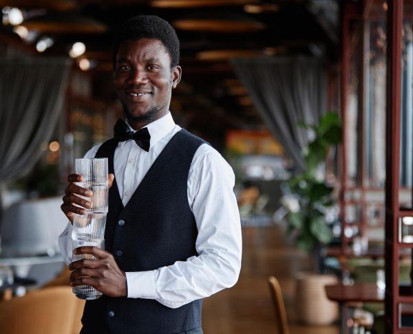 waist-up-portrait-young-black-man-as-elegant-server-holding-glasses-smiling-camera-luxury-restaurant-copy-space